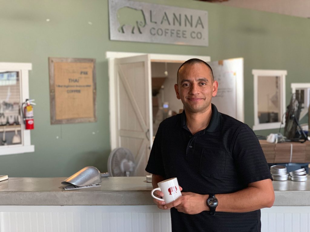 A man in a black shirt stands behind a counter holding a white mug, smiling at the camera. Behind him is a sign that reads “Lanna Coffee Co.” and a rustic interior with shelves and equipment.