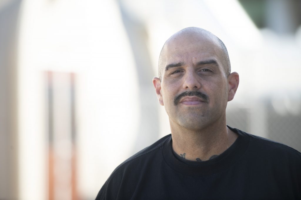 A man with a shaved head and mustache, wearing a black shirt, stands outdoors in front of a blurred light-colored background, looking directly at the camera with a neutral expression.