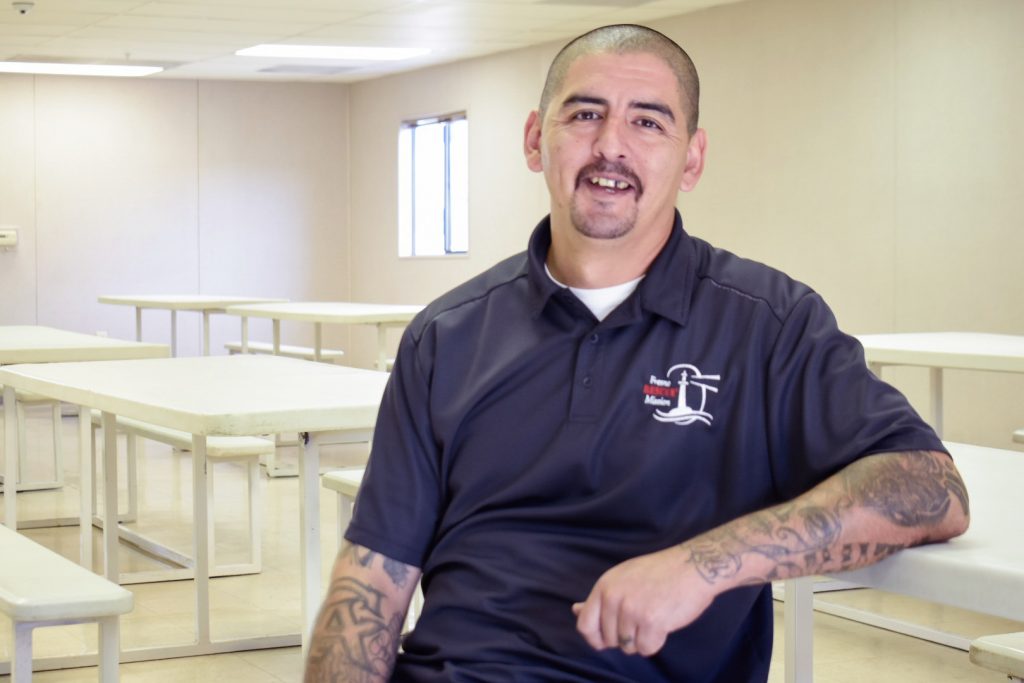 A man with tattoos, short hair, and a goatee, wearing a navy polo shirt with a logo, sits and smiles in a bright, empty room with rows of white tables.