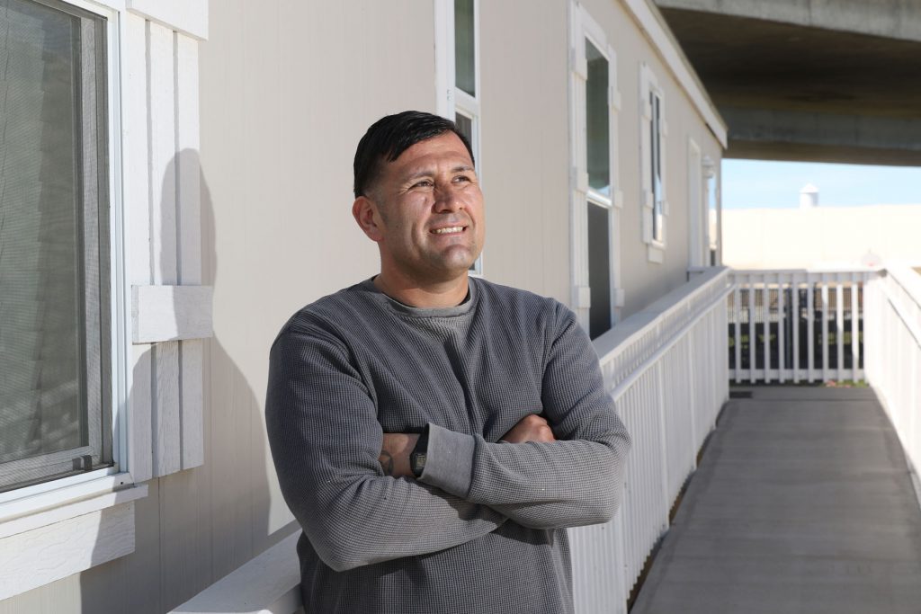 A man with short dark hair, wearing a gray long-sleeve shirt, stands outside a light-colored building with his arms crossed, smiling and looking to the side.