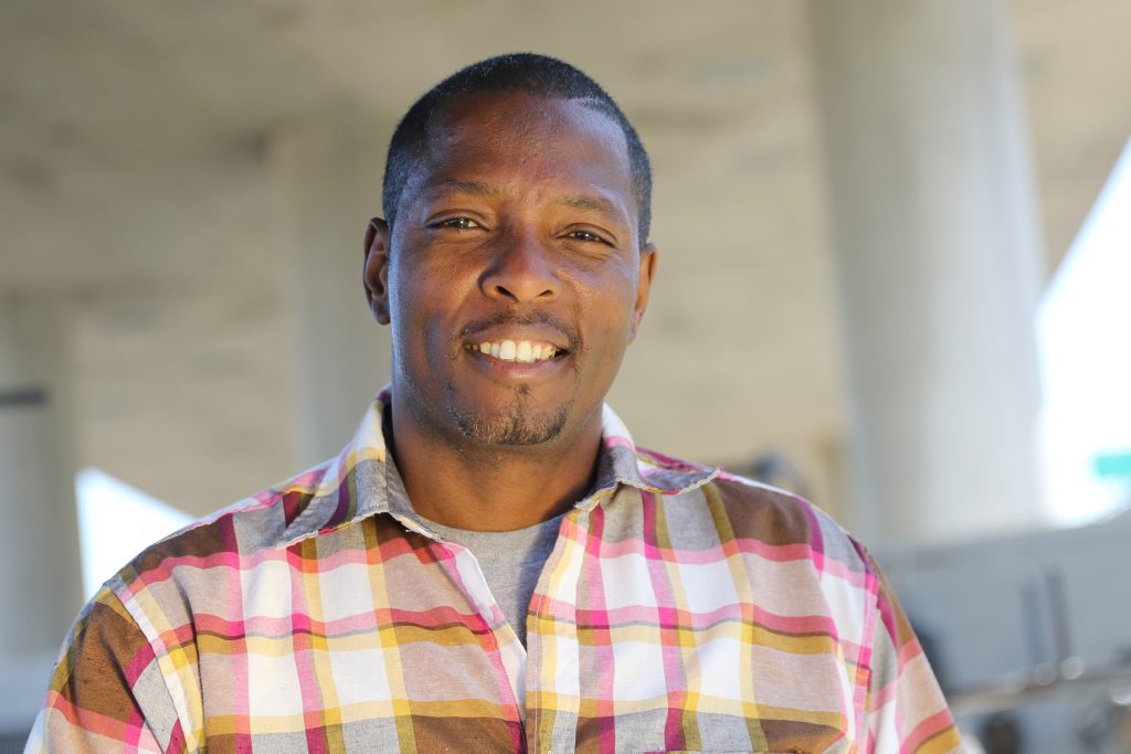 A man wearing a plaid shirt smiles at the camera. The background is blurred, showing large concrete pillars and soft daylight.