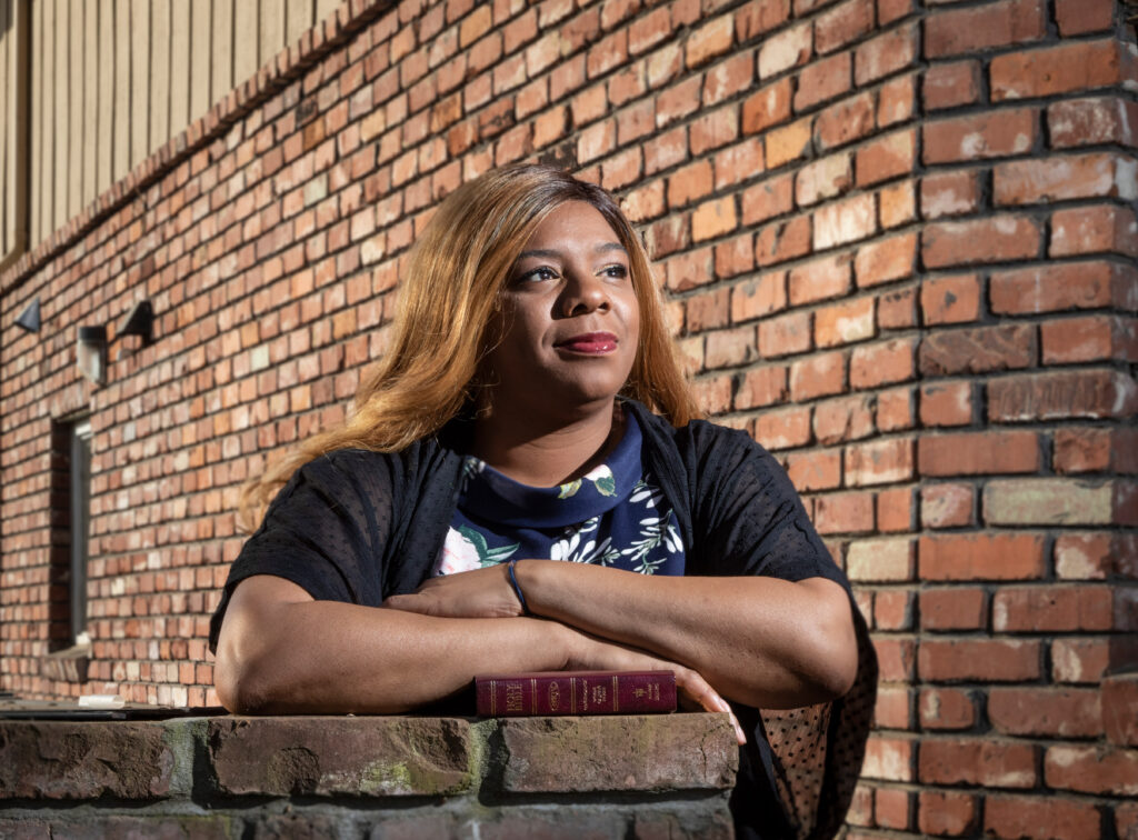 A woman with long hair leans on a brick wall, arms crossed over a small book, gazing thoughtfully into the distance. She wears a floral top and dark cardigan. The background is a sunlit brick building.