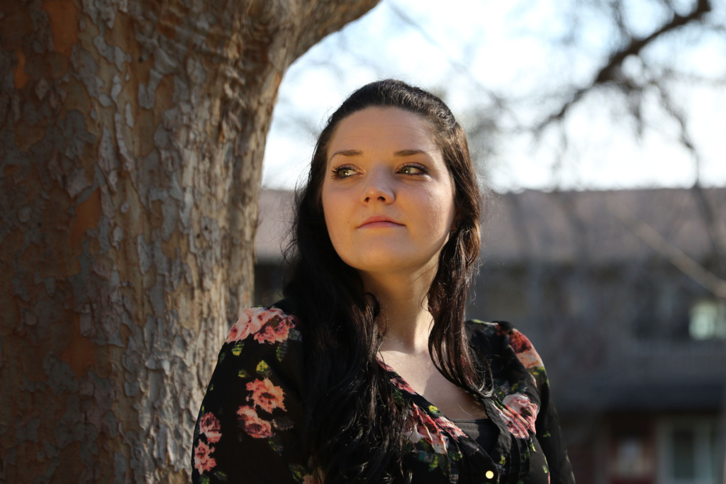 A woman with long dark hair, wearing a black floral top, stands outdoors beside a tree, looking thoughtfully into the distance with soft sunlight illuminating her face.