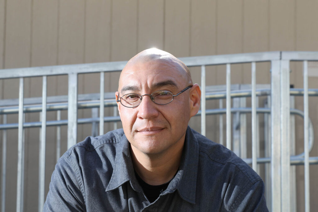 A man with glasses and a shaved head is sitting outdoors in front of a metal railing, wearing a dark button-up shirt and smiling slightly at the camera.