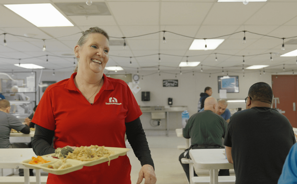 A smiling woman in a red shirt holds a tray of food in a cafeteria with several people sitting at white tables eating meals under string lights.