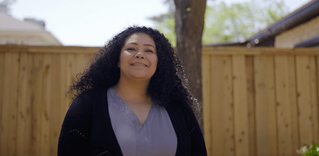 A woman with long curly hair smiles outdoors, standing in front of a wooden fence and trees on a sunny day.
