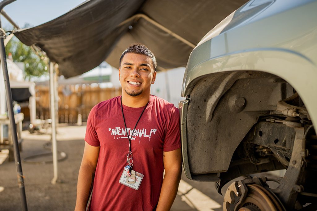 A young man wearing a red Intentional t-shirt and a lanyard smiles while standing next to a car with its wheel removed in an outdoor automotive work area.