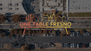 Aerial view of a parking lot and people gathering outside buildings, overlaid with a colorful Fresno skyline illustration and the text “One Table Fresno: One Purpose. One Cause. One Table.”.