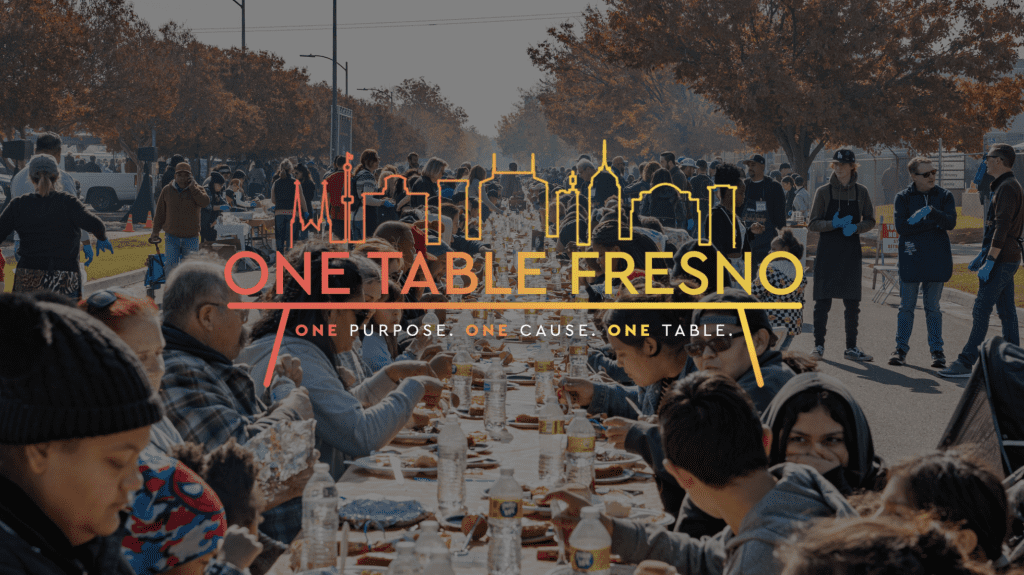People gather outdoors at a long table for a communal meal. The text ONE TABLE FRESNO: One Purpose, One Cause, One Table with a city skyline is overlaid on the image. Trees with autumn leaves line the street.