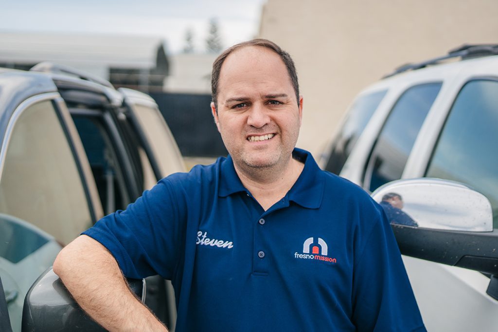 A man wearing a blue Fresno Mission polo shirt and name tag “Steven” smiles while standing between two parked vehicles.