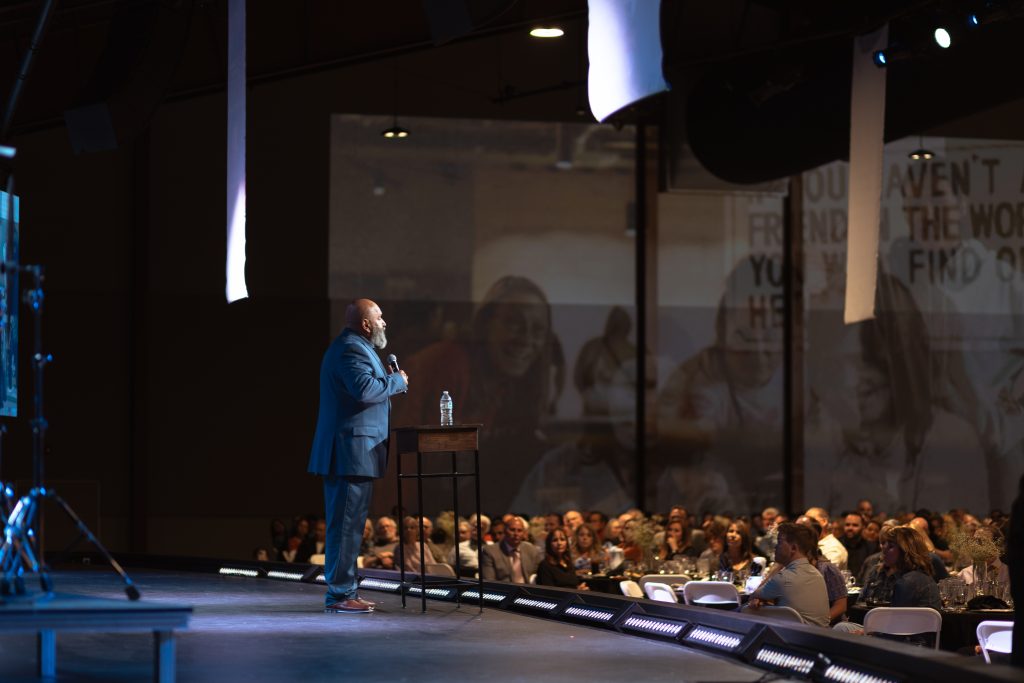 A man in a suit speaks on stage to a seated audience in a large, dimly lit auditorium. A small table with water is beside him, and a large projection is visible behind him.