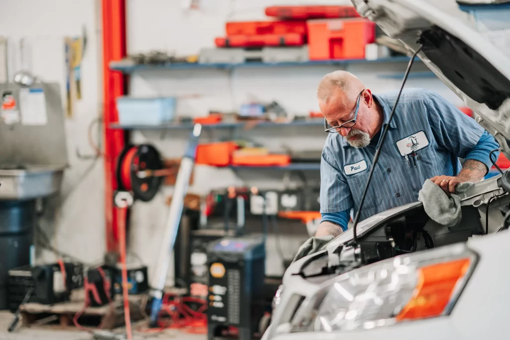 A mechanic with glasses and a blue shirt works under the hood of a white car in a garage, surrounded by tools and equipment on shelves in the background.