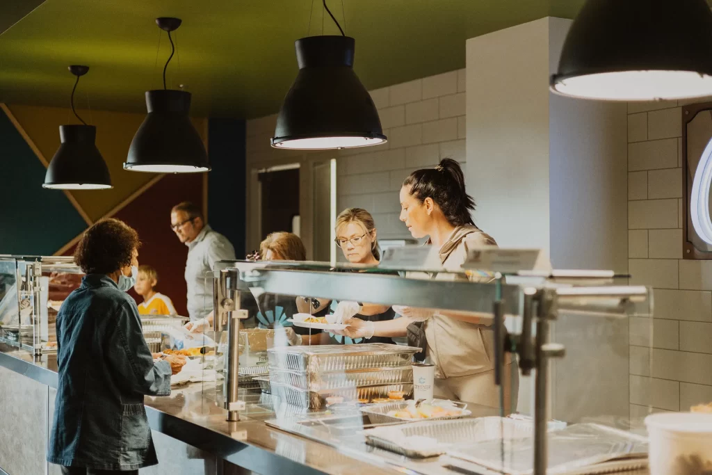People serving food behind a cafeteria counter to individuals on the other side; overhead lights illuminate the food and the servers, and several people are visible waiting in line.