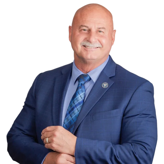 A smiling older man with a bald head and mustache wears a blue suit, blue patterned tie, and a pin on his lapel. He stands with one hand holding his other wrist, posing against a plain background.