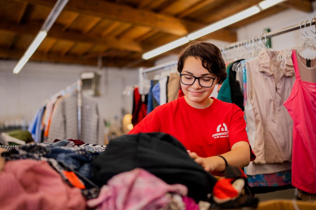 A person wearing glasses and a red shirt sorts through a pile of clothes in a thrift store or donation center, surrounded by racks of hanging clothing under a wooden ceiling.